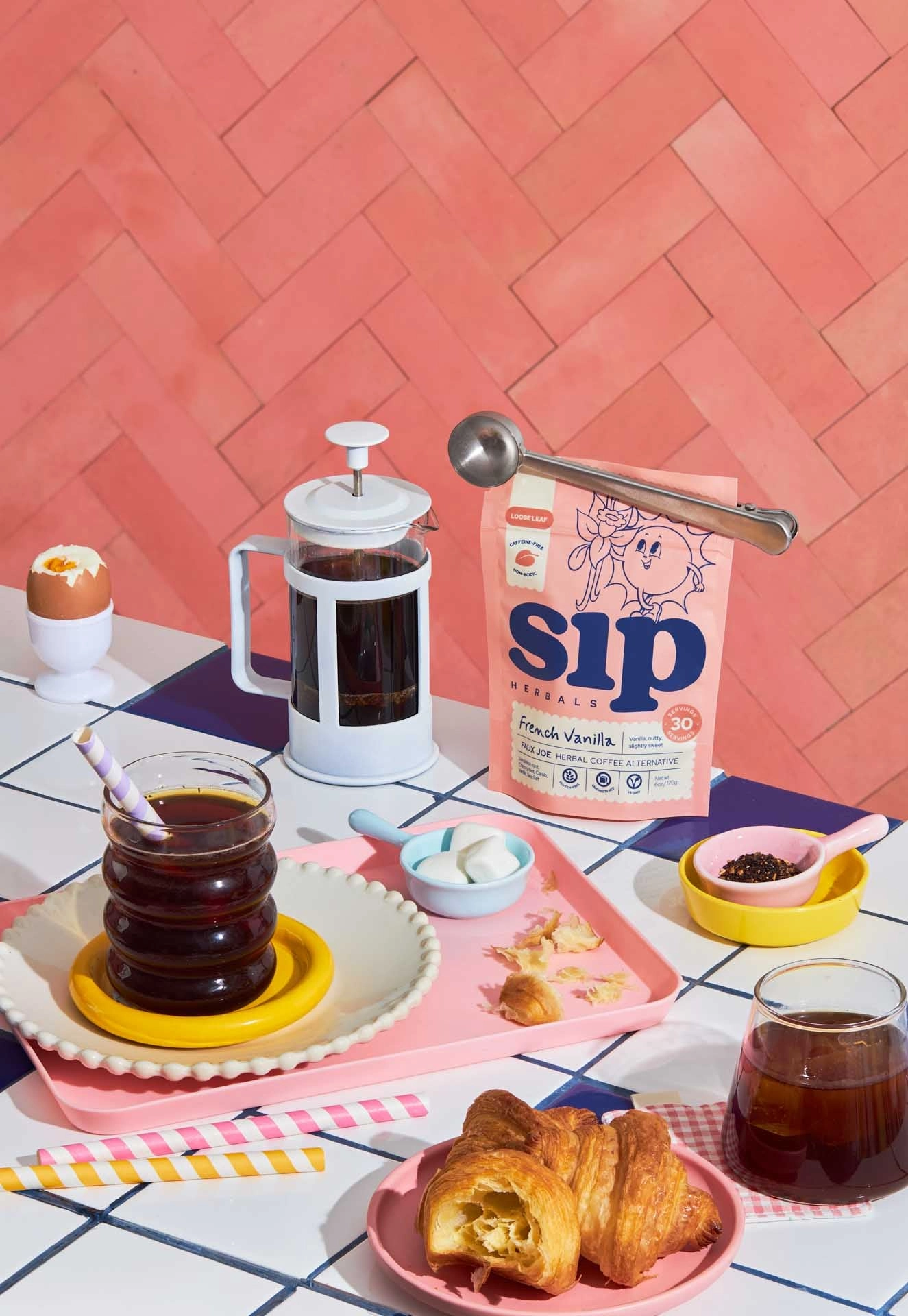 Sip Herbals French Vanilla herbal coffee alternative bag styled on a colorful tabletop with a white French press, an iced drink, croissants and a metal scoop